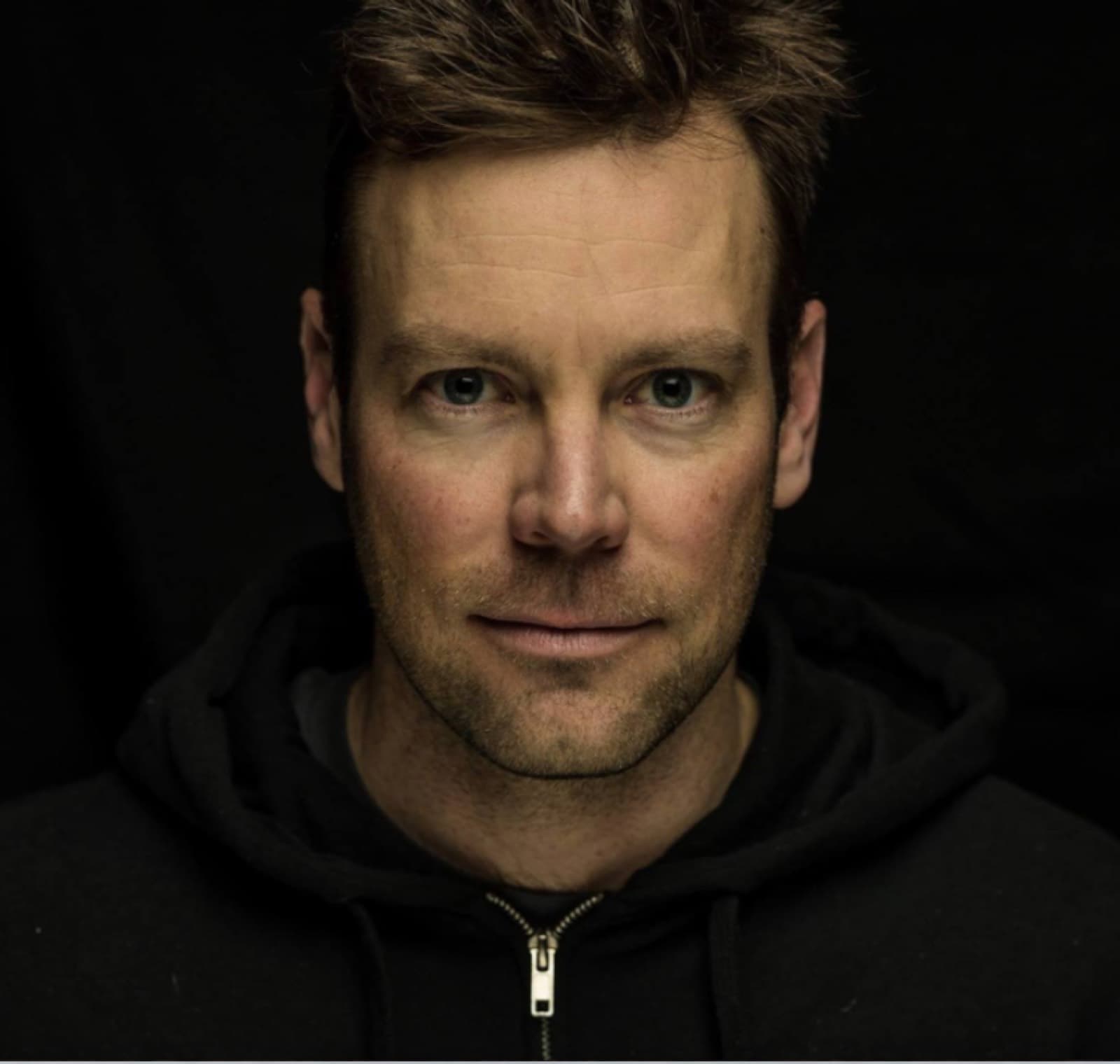 Head-and-shoulders portrait of a man in a black hooded sweatshirt against a dark background