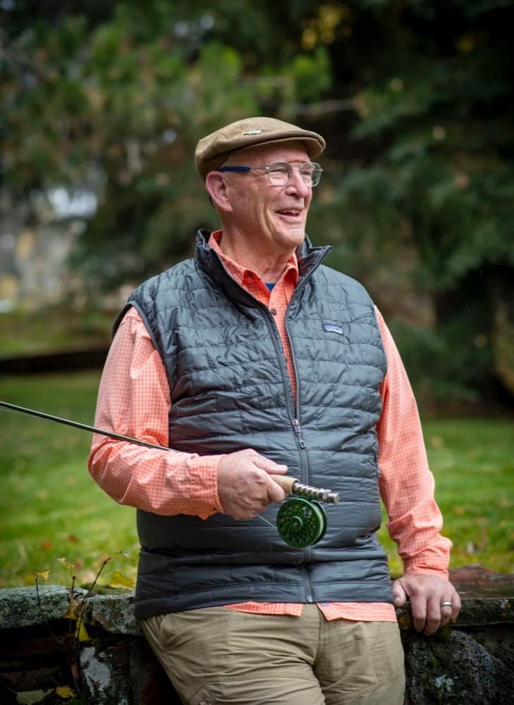 An angler in a flat cap and glasses, smiling, holding a fly rod and reel beside a stone wall