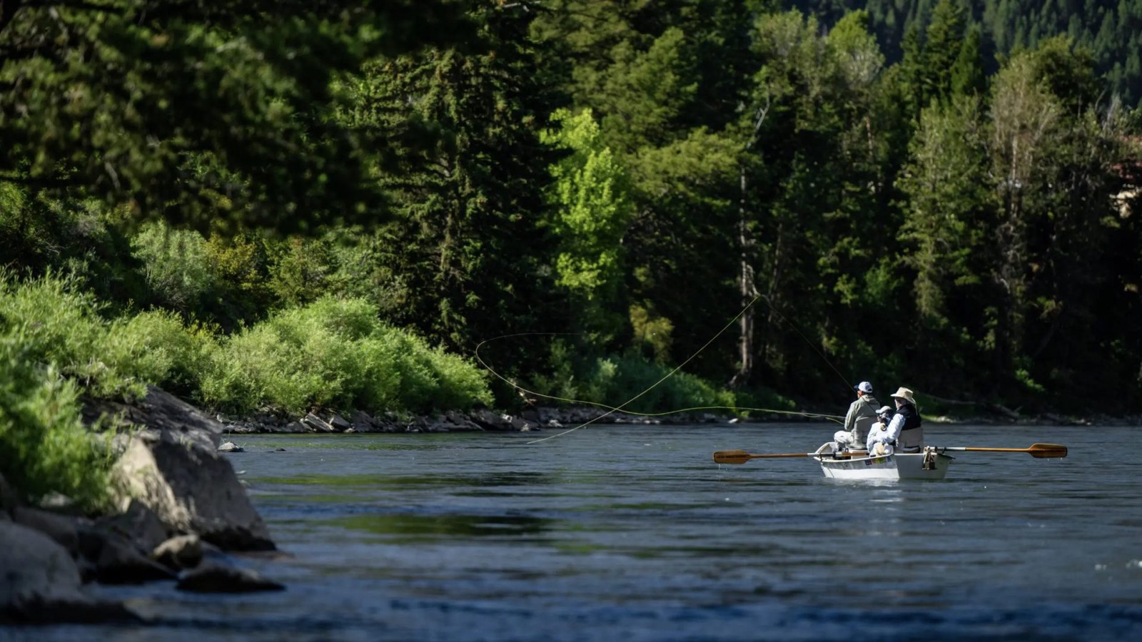 Anglers fly casting from a white drift boat on a river below a forested bank