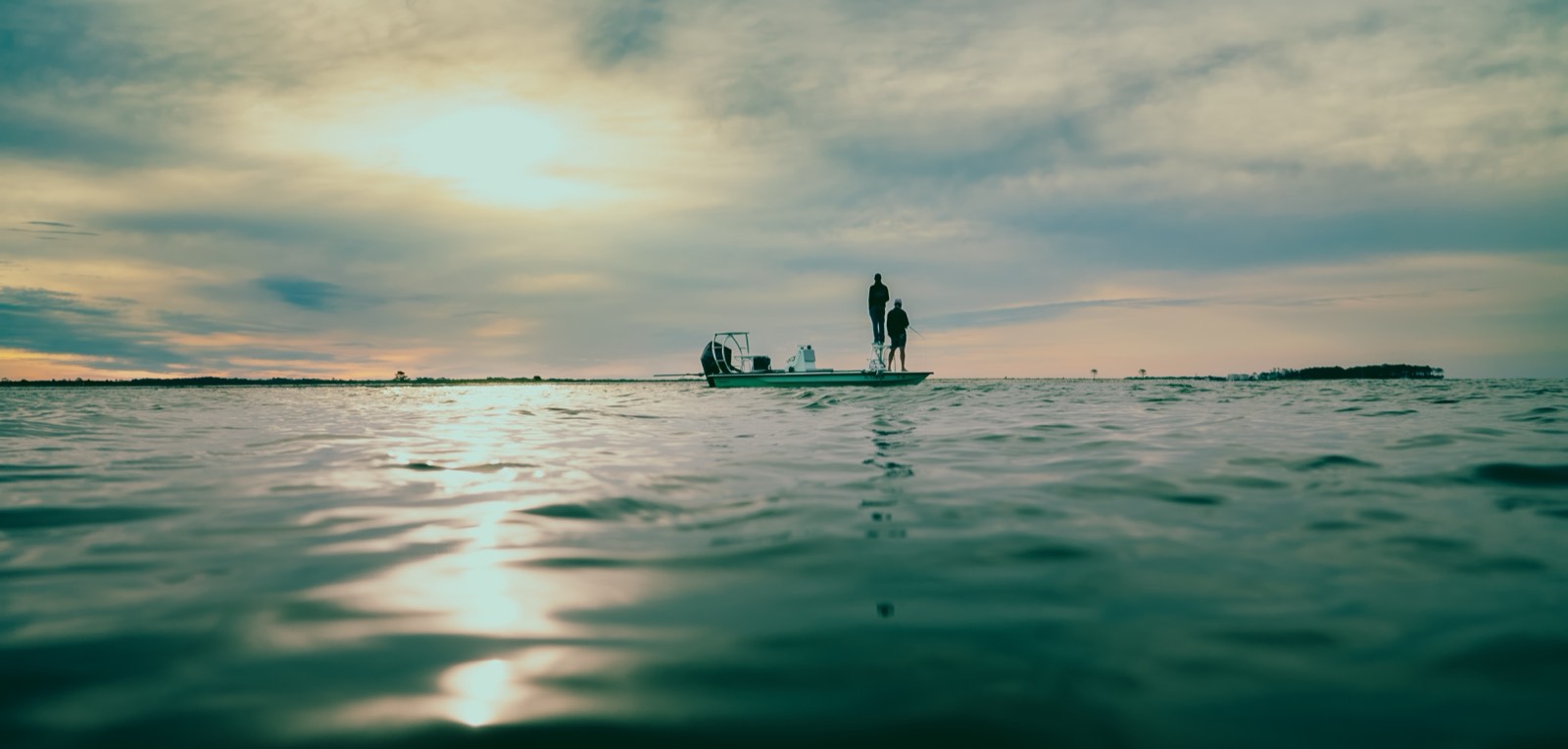 Two anglers standing on a flats boat on open water under a cloudy dusk sky